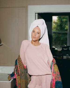 A model in a hair towel stands in a vintage bathroom holding a colorful patchwork kimono over her arm, wearing a soft pink waffle-knit PJ set — the whole thing reads like the best kind of slow morning.