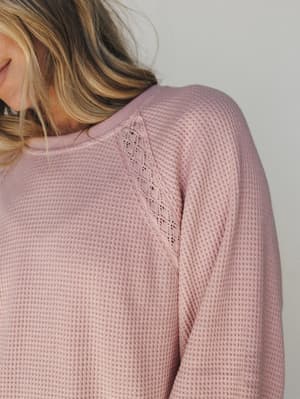 A close-up detail shot of a dusty pink waffle-knit top with a delicate lace inset along the raglan seam — the kind of cozy piece that makes staying in feel intentional.