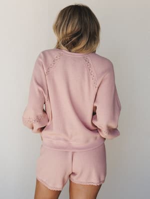 A matching waffle-knit top and shorts set in soft blush pink, shot from behind to highlight the delicate lace trim detailing along the raglan seams and sleeve cuffs — cozy and pretty in equal measure.