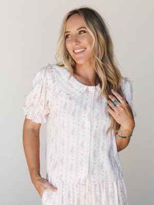 A smiling model in a soft floral two-piece set with a peter pan collar and puff sleeves — the kind of thing you'd wear on a slow morning and somehow never want to take off.