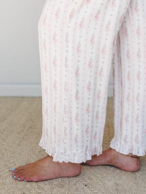 A close-up of flowy floral pajama pants with a delicate ruffle hem, shot at ankle level on a jute rug — soft, dreamy, and made for slow mornings.