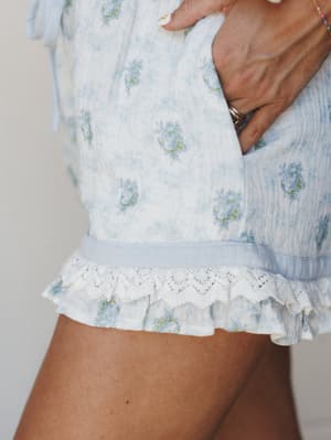 A close-up detail shot of floral print shorts with a ruffled lace hem and elastic waistband, shot in soft natural light — the kind of thing you'd want to wake up in on a slow Sunday.