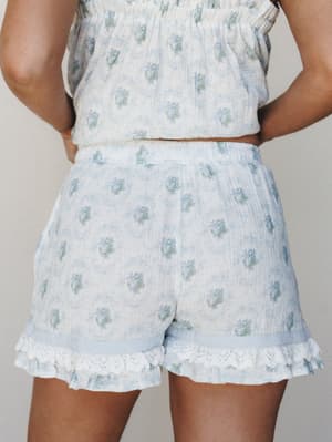 A close-up back view of a matching floral short set in soft light blue and cream — the shorts feature a ruffle lace hem and elasticated waist, paired with a matching smocked crop top, shot against a neutral studio backdrop.