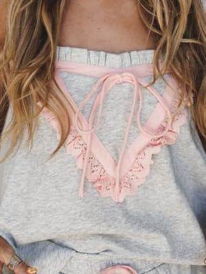 A close-up detail shot of a soft grey sweatshirt-style top with blush pink lace trim, ruffle collar, and a delicate ribbon tie at the neckline — cozy and feminine in equal measure.