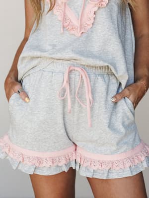 A cropped mid-shot of a heathered grey matching short set with soft pink lace ruffle trim at the hem and neckline — the kind of set that makes staying in feel like a whole vibe.