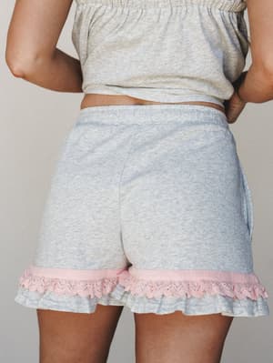 A close-up back view of heathered grey pull-on shorts with a sweet pink ruffle and lace trim at the hem, styled as part of a matching two-piece set — soft, cozy, and just feminine enough.