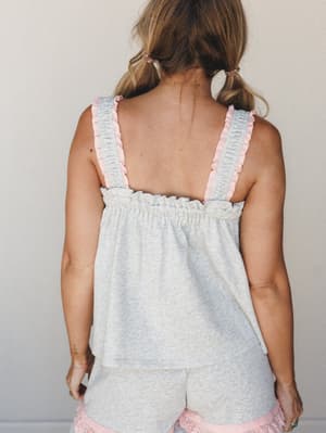 A back-view shot of a heathered grey cami top with pink ruffle-trimmed straps and a gathered empire waist, paired with matching grey and pink shorts — soft, sweet, and made for slow mornings.