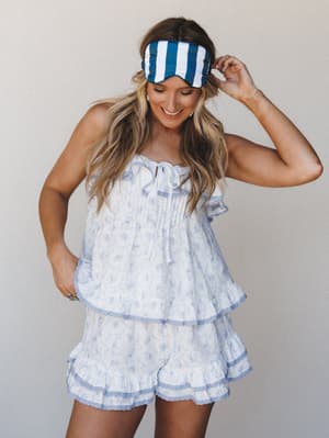 A smiling model wears a soft floral pajama set in white and light blue — ruffled cami top and matching shorts — with a blue striped sleep mask pushed up on her head, giving the whole look an effortlessly playful, 'good morning' energy.