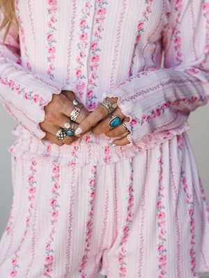 A close-up detail shot of a pink floral ribbed PJ short set, hands folded at the waist and layered with silver and turquoise rings — the kind of cozy-meets-collected moment that makes loungewear feel intentional.