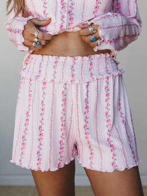 A close-up of a matching floral short set in soft pink — the smocked, ruffled waistband and lettuce-edge hem give it that sweet, lived-in feel, styled with stacked silver rings for just the right amount of personality.