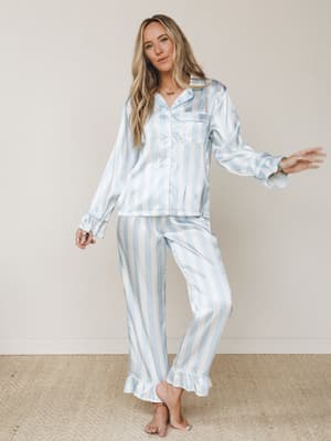 A satin stripe pajama set in soft sky blue and cream, styled on a barefoot model with a playful, easy stance — the kind of set that makes staying in feel like a whole mood.