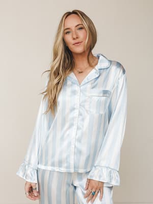 A satin striped pajama set in powder blue and white, styled with layered gold necklaces and stacked rings — the kind of set that makes staying in feel like a whole mood.