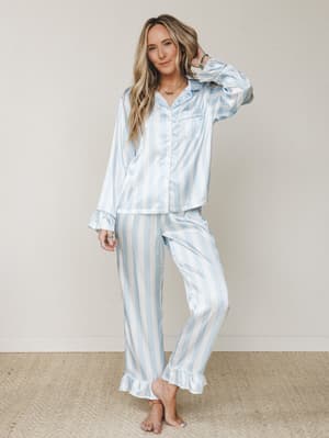 A satin stripe pajama set in soft sky blue and white, styled on a barefoot model with layered jewelry — the kind of set that makes staying in feel like the better plan.