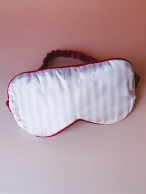 A satin sleep mask in soft blush with tonal stripe texture and deep raspberry piping, laid flat against a dusty rose background — the kind of thing that makes going to bed feel like a small ritual.