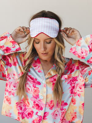 A model with loose pigtail braids adjusts a pink gingham sleep mask on her head while wearing a bold satin floral pajama top — soft, dreamy, and somehow the most put-together version of staying in.