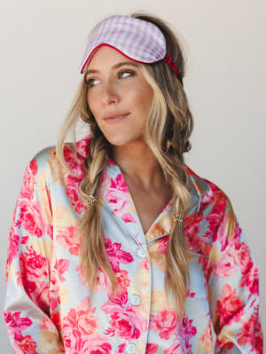A dreamy, playful shot of a model wearing a pink gingham sleep mask pushed up on her head like a headband, paired with a satin floral pajama top in full bloom — the kind of look that makes staying in feel like a whole occasion.