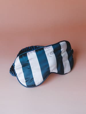 A satin sleep mask in bold navy and white stripes sits against a warm dusty rose background — the kind of thing that makes a Sunday morning feel intentional.