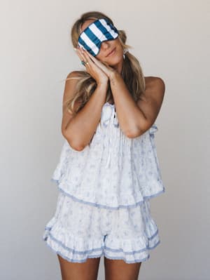 A model wearing a flowy floral cami top and matching ruffle shorts poses with hands tucked under her cheek, a teal and white striped satin sleep mask covering her eyes — the whole look reads like a dreamy Saturday morning with nowhere to be.