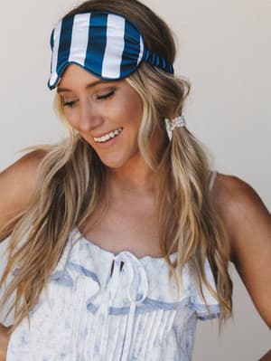 A smiling model wears a navy and white striped satin sleep mask pushed up on her forehead like a headband, paired with a soft blue floral ruffle-trim camisole and a pearl hair tie — the whole look feels like a really good morning.