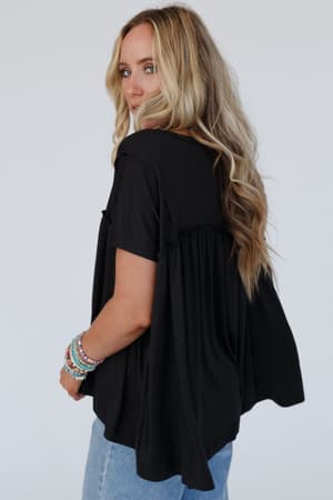 A woman with long, wavy blonde hair wears the Sunday Morning Babydoll Top - Black with light blue jeans and colorful beaded bracelets. She looks over her shoulder against a plain white background.