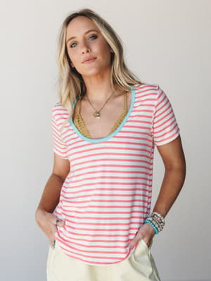 A relaxed coral stripe tee with a playful turquoise scoop neck trim, styled with layered boho jewelry and pale yellow pants — the kind of effortless summer outfit that looks like you tried just the right amount.