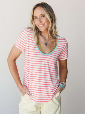 A relaxed striped scoop-neck tee in coral and white gets a playful twist with a contrasting turquoise neckline trim, styled with soft yellow pants and layered boho jewelry for an easy, warm-weather feel.