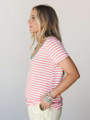 A side profile shot of a model in a coral and white striped scoop neck tee tucked loosely into cream pants, styled with stacked turquoise and neutral bracelets — the kind of easy, happy outfit that makes a Tuesday feel like a vacation day.