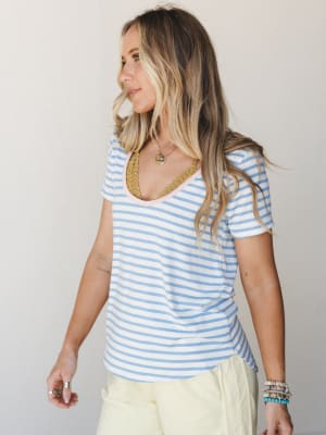 A relaxed striped scoop-neck tee in soft sky blue and white, styled with pale yellow bottoms and layered gold and beaded jewelry — the kind of effortless everyday look that just works.