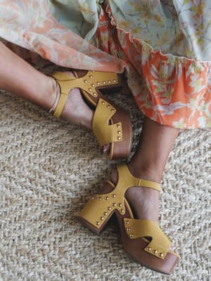 A close-up of mustard yellow studded block heel sandals paired with a flowy floral tiered skirt, shot from above on a woven jute rug — the kind of detail shot that makes you wish you were already running late to something fun.