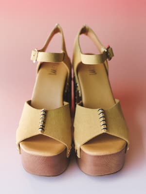 A pair of yellow suede platform heel sandals with a wood-look sole, ankle strap, and whipstitch hardware detail, shot flat against a soft pink gradient background.