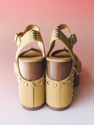 A pair of mustard yellow suede platform sandals shot from behind against a soft pink backdrop — the wood block heel and silver stud detailing give them that perfect retro-boho attitude.