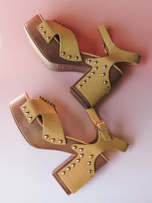 A pair of mustard suede platform clog sandals with gold stud detailing and ankle buckle straps, casually scattered on a blush pink surface — the kind of shoes that make a whole outfit feel intentional.