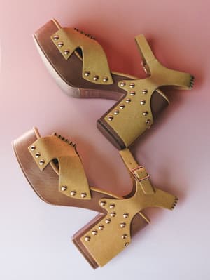 A pair of mustard suede platform clog sandals with gold stud detailing and ankle buckle straps, casually scattered on a blush pink surface — the kind of shoes that make a whole outfit feel intentional.