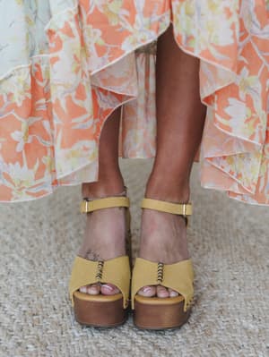 A close-up detail shot of mustard yellow platform clog sandals paired with a flowy floral ruffle skirt in peach and cream — the kind of footwear that makes an outfit feel both grounded and a little wild.