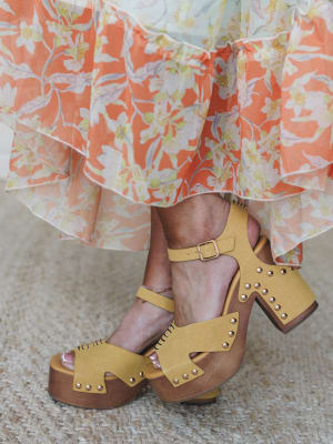 A close-up of mustard yellow platform clog sandals with gold stud detailing and ankle straps, paired with a flowy coral floral tiered skirt — the kind of outfit that feels like a warm afternoon even when it isn't.