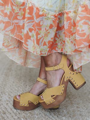 A close-up of mustard yellow platform clog sandals with gold stud detailing and ankle straps, paired with a flowy coral floral tiered skirt — the kind of outfit that feels like a warm afternoon even when it isn't.
