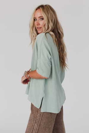 A woman with long blonde hair wears The Nest On The Go V Neck Ribbed Essential Tee in sage from Three Bird Nest, brown pants, and colorful beaded bracelets, looking over her shoulder against a plain light background.