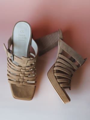 A pair of khaki suede strappy heeled sandals laid flat against a soft rose-to-white gradient background — woven toe straps, a buckle ankle strap, and a ribbed block heel give them that perfect mix of boho structure and easy summer dressing.