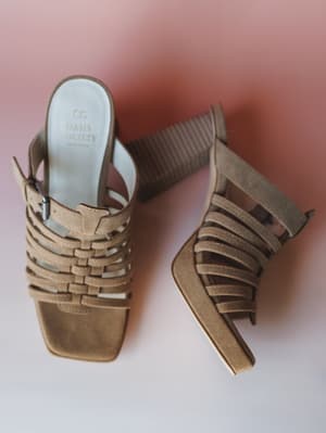 A pair of khaki suede strappy heeled sandals laid flat against a soft rose-to-white gradient background — woven toe straps, a buckle ankle strap, and a ribbed block heel give them that perfect mix of boho structure and easy summer dressing.