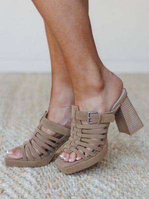A close-up of strappy khaki suede platform mules with a chunky stacked block heel, shot at ankle level on a jute rug — the kind of shoe that does the heavy lifting for an otherwise easy outfit.