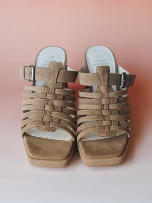 A pair of khaki suede strappy platform heels shot from the front against a soft blush gradient background — woven cage straps and silver buckle hardware give them that effortless, slightly retro-boho energy.