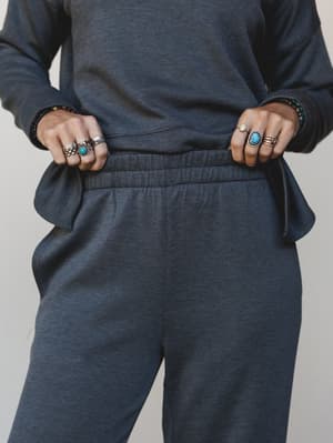 A close-up mid-shot of a matching charcoal knit set — the model's hands grip the elastic waistband of the relaxed pants, showing off a full stack of silver and turquoise rings and beaded bracelets that do most of the talking.