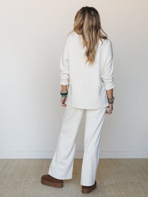 A matching pullover-and-wide-leg-pant set in heathered vanilla worn from the back, styled with stacked beaded bracelets and brown platform clogs — the kind of effortless monochromatic outfit that makes a Saturday feel intentional.