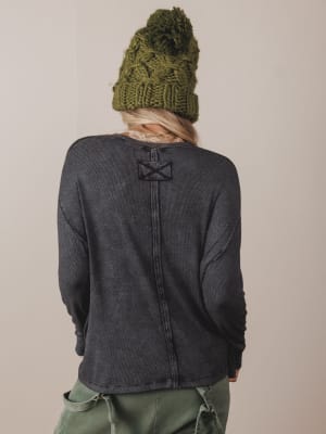 Person wearing a dark gray long-sleeve shirt with a logo on the back, green pants, and a green knit beanie against a beige background.