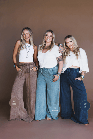 Three women smile together against a brown backdrop in Relaxing Robin Wide Leg Pant - New Teal with drawstring waists and white tops. Heights and pant sizes: 5’8” Small, 5’6” Medium, 5’3” XL.