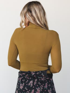 Shot from the back, this olive gold turtleneck bodysuit is fitted and long-sleeved, paired with a dark floral skirt — the kind of put-together layering that feels intentional without trying too hard.