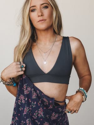 A close-cropped model shot of a charcoal bralette layered with stacked silver necklaces and an armful of boho bracelets — the floral skirt pulled open below gives it that effortless, half-dressed energy.