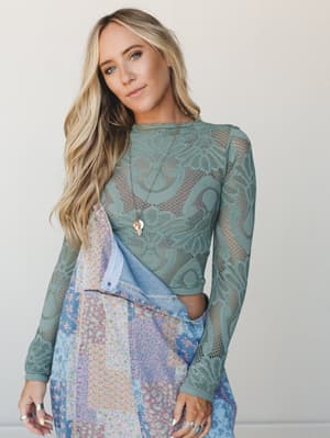 A long-sleeve sage lace net crop top layered over a patchwork floral skirt and denim, styled with layered gold necklaces and stacked rings — the kind of outfit that looks like it came together effortlessly but absolutely did not.