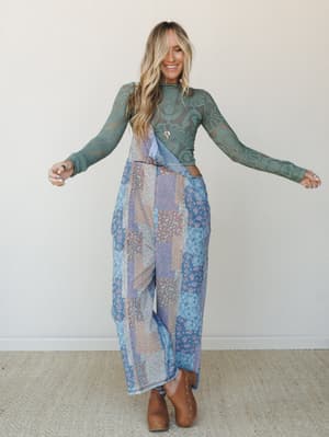 A smiling model with arms outstretched wears a sage green lace long-sleeve crop top paired with wide-leg patchwork print pants and tan studded clogs — the whole look feels like a bohemian daydream you'd actually wear.