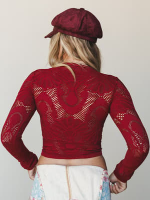Shot from behind, she's wearing a deep merlot crochet-lace crop top with long sleeves and an open mesh pattern, paired with a matching suede baker boy cap and a floral patchwork skirt peeking at the hem — the whole look has a cool, vintage-boho confi